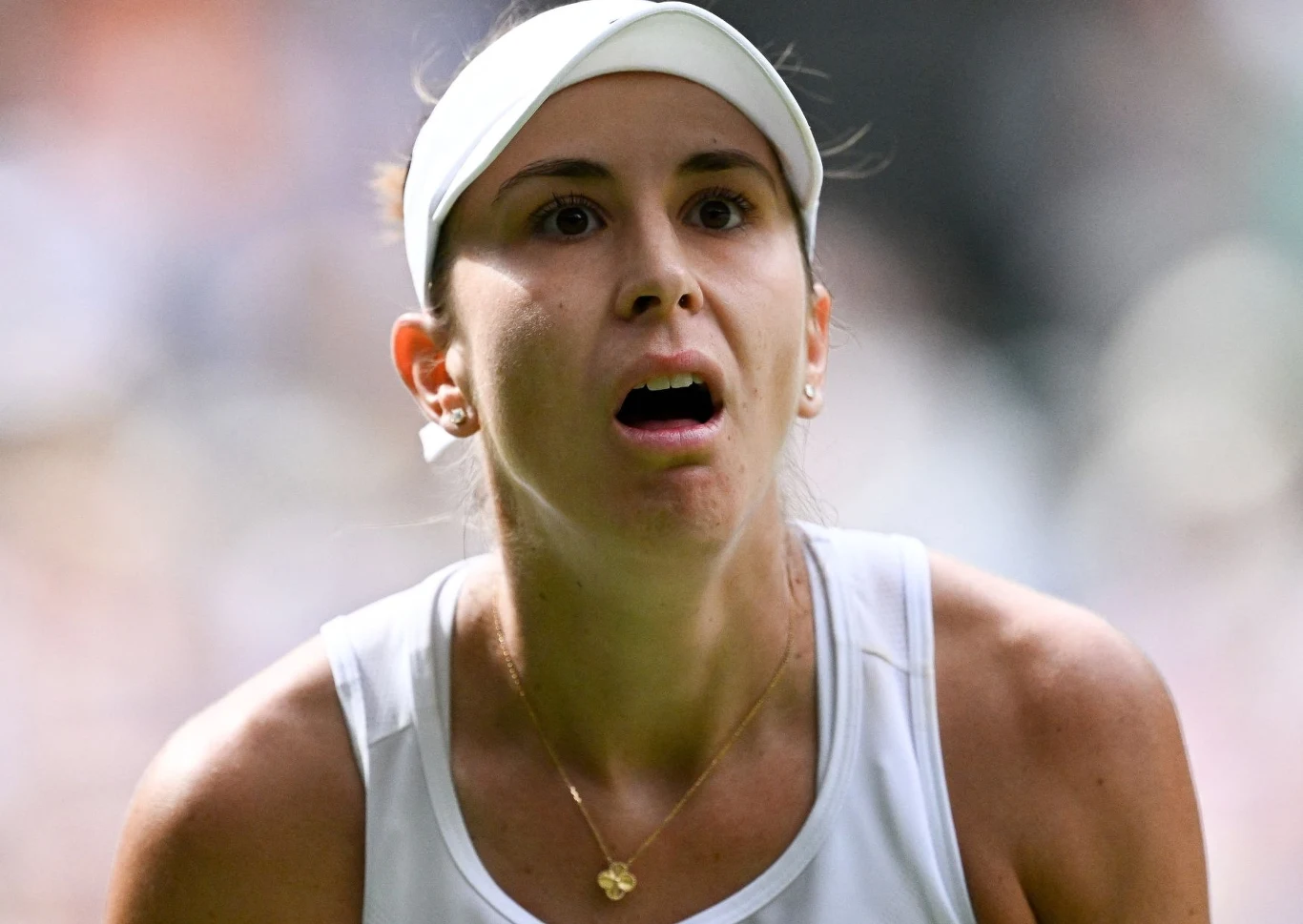 Zbliżenie twarzy kobiety o jasnych włosach spiętych opaską, ubranej w biały sportowy strój, z intensywnym wyrazem skupienia i otwartymi ustami, tło rozmyte.