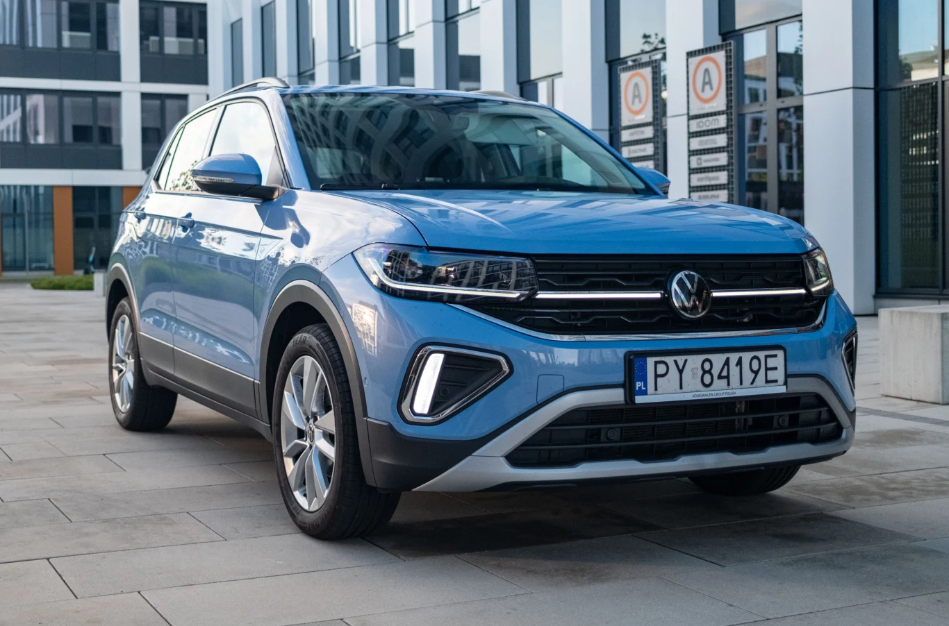 T-Cross z powodzeniem może służyć jako auto rodzinne, pod warunkiem, że nie musimy wozić już dużego wózka na wakacje. Niebieski samochód marki Volkswagen zaparkowany na tle nowoczesnego biurowca, wyeksponowany przód pojazdu, dobrze widoczne reflektory, tablica rejestracyjna oraz przeszklona fasada budynku w tle.