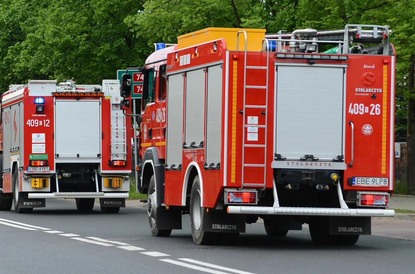 Czy można wyprzedzić kolumnę straży pożarnej? Dwa czerwone wozy strażackie jadące ulicą w stronę sygnalizacji świetlnej, otoczone drzewami i zielenią, z widocznymi numerami operacyjnymi i oznaczeniami jednostki.