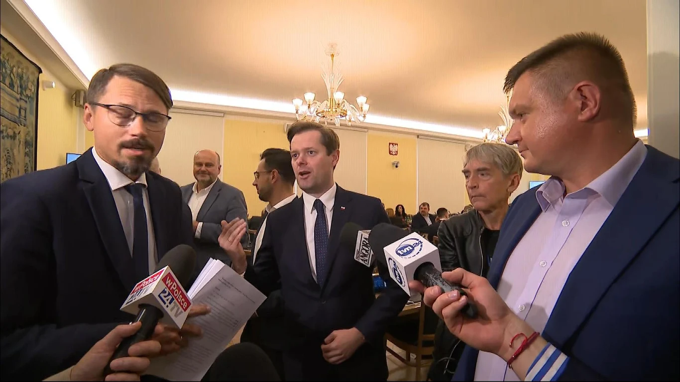 Kłótnia na sejmowej komisji finansów publicznych Grupa mężczyzn w garniturach rozmawia z dziennikarzami, wokół nich widać mikrofony z logotypami stacji telewizyjnych, w tle elegancko urządzona sala z żyrandolami i elementami godła państwowego.