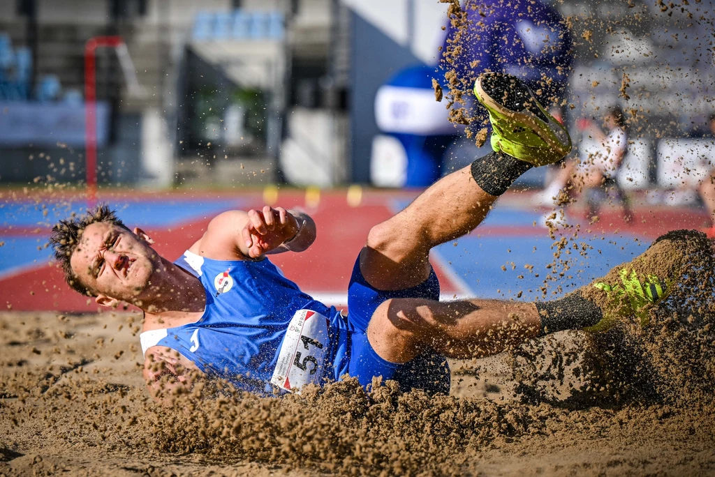 Bartosz Sienkiewicz Zawodnik uprawiający lekkoatletykę w niebieskim stroju wpada w piasek podczas skoku w dal, wokół unoszą się drobinki piasku, tło rozmyte, skupione na sportowcu.