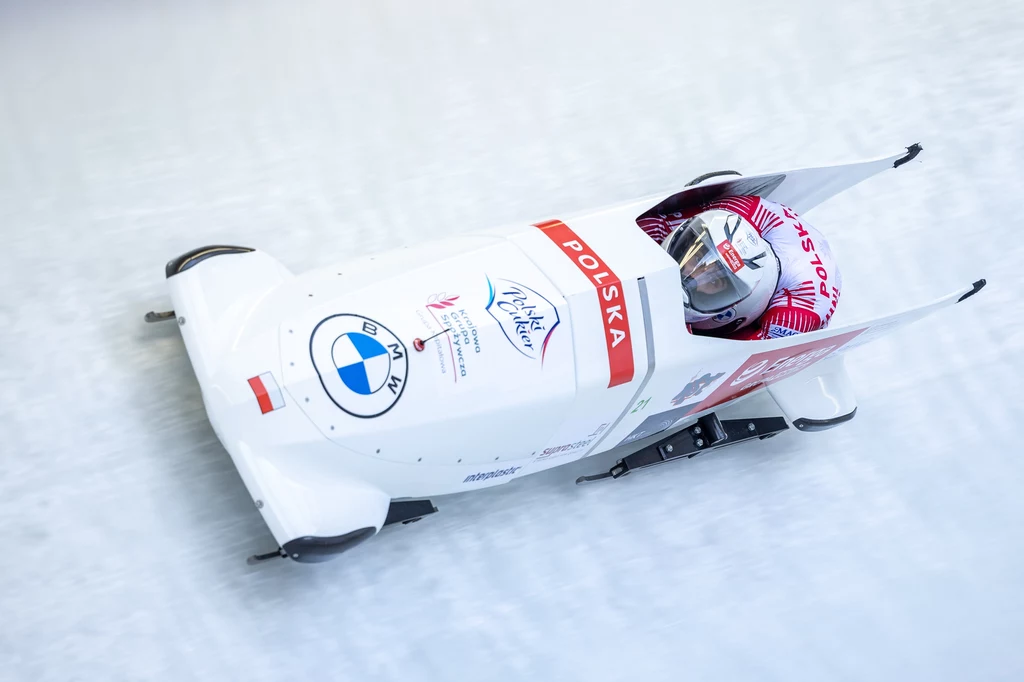 Bartosz Sienkiewicz w rywalizacji w bobslejach Bobslej z polską flagą oraz logotypami sponsorów sunący po torze lodowym, wewnątrz zawodnik ubrany w biało-czerwony kask i kombinezon.