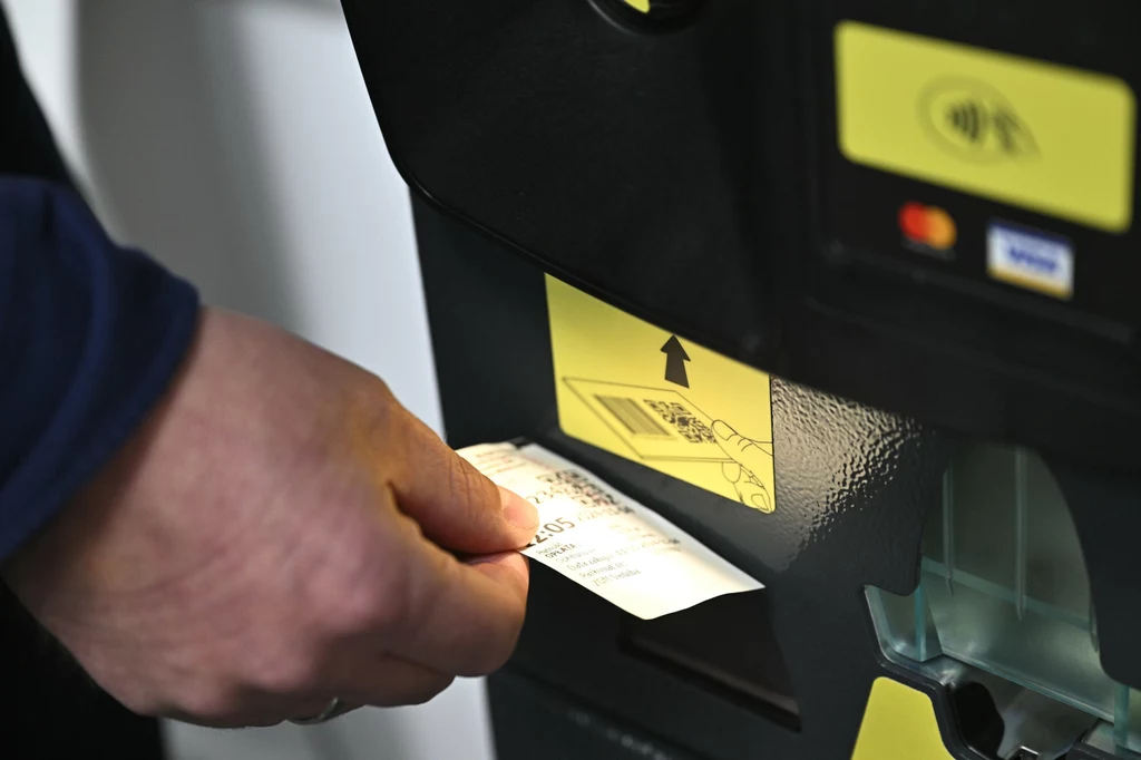 Niektóre z nowoczesnych parkomatów umożliwiają płatność wyłącznie bezgotówkową. Dłoń wsuwająca bilet parkingowy do automatu, wyraźnie widoczna instrukcja korzystania z maszyny oraz kolorowe naklejki informacyjne.