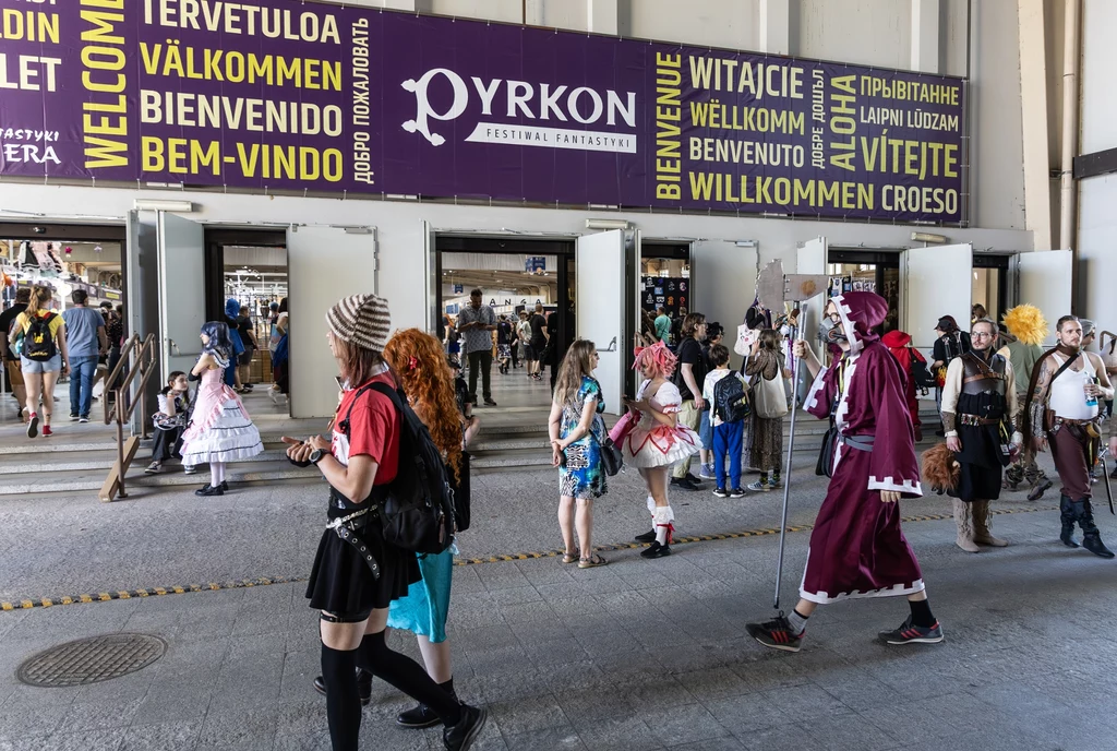W 2025 roku w Pyrkonie wzięła udział rekordowa liczba uczestników Ludzie w kolorowych, fantazyjnych kostiumach przechodzą przez wejście budynku, nad którym widnieją wielojęzyczne powitania oraz baner informujący o festiwalu Pyrkon. Przestrzeń wypełniona jest uczestnikami konwentu, z których niektórzy wcielają się w p...