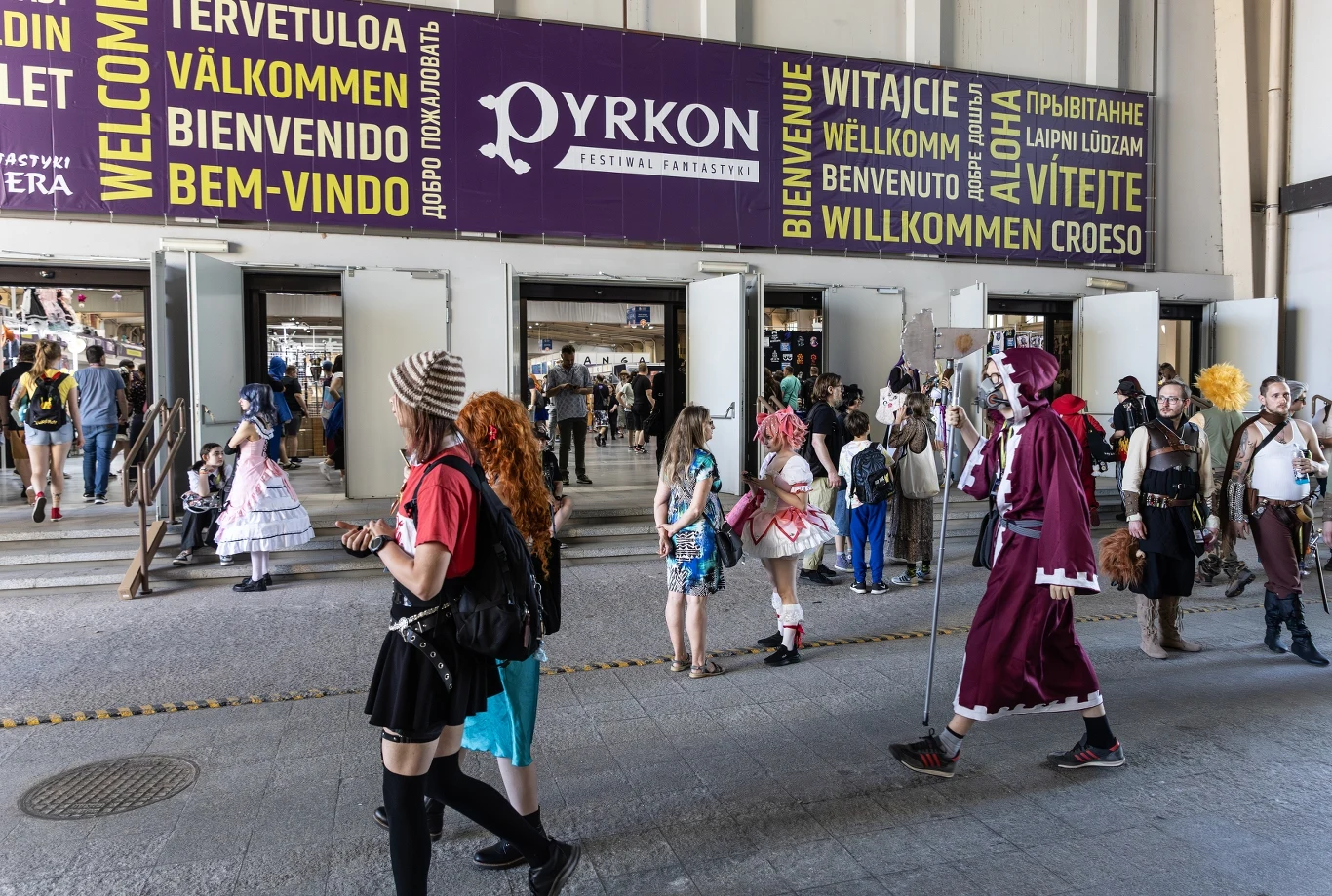 Ludzie w kolorowych, fantazyjnych kostiumach przechodzą przez wejście budynku, nad którym widnieją wielojęzyczne powitania oraz baner informujący o festiwalu Pyrkon. Przestrzeń wypełniona jest uczestnikami konwentu, z których niektórzy wcielają się w p...