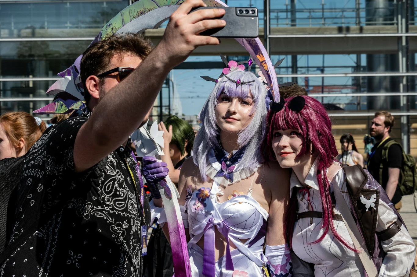 Grupa osób robiących selfie podczas konwentu, dwie kobiety ubrane w barwne stroje cosplay inspirowane postaciami z kultury japońskiej, w tle widać przechodniów oraz nowoczesny budynek.
