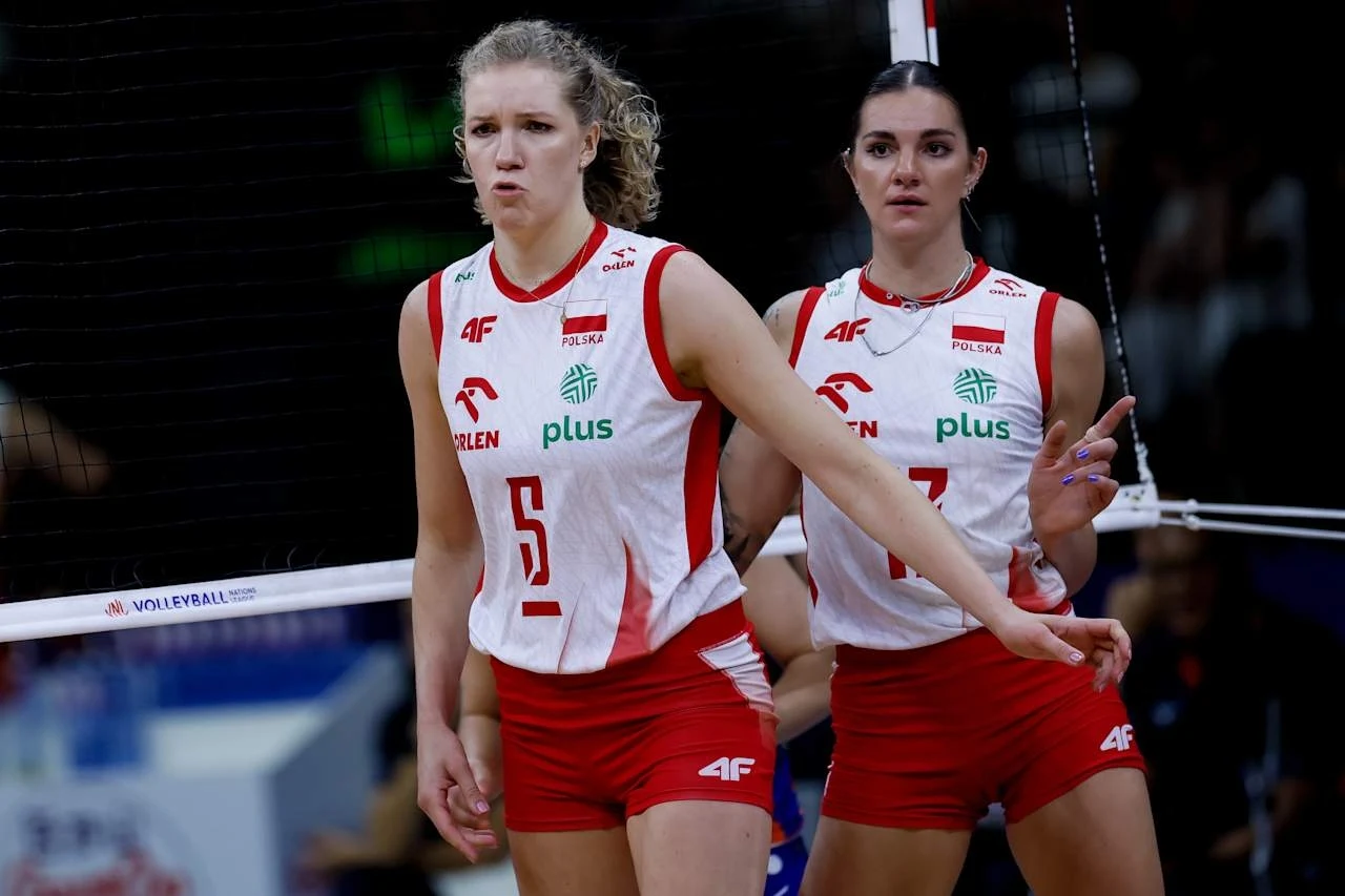 Malwina Smarzek (z prawej). Obok Agnieszka Korneluk Dwie siatkarki polskiej reprezentacji w biało-czerwonych strojach, stojące blisko siatki na hali sportowej, skupione na grze, w tle widoczny fragment boiska oraz siatka.