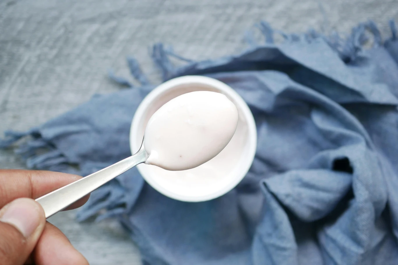 Jogurt na zdrowe jelita Łyżeczka z jogurtem trzymana nad białym kubeczkiem ustawionym na niebieskiej tkaninie.