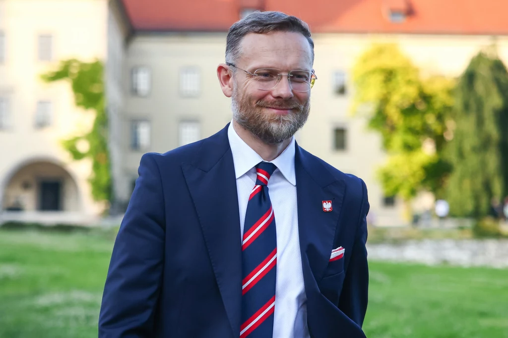 Poseł PiS Zbigniew Bogucki Mężczyzna w eleganckim garniturze i krawacie w prążki stoi na trawie przed jasnym budynkiem z czerwonym dachem, uśmiecha się, ma okulary i brodę.