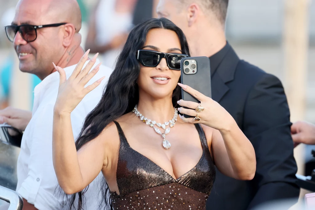 Kim Kardashian uwielbia drogą biżuterię, a założenie tej należącej do Elizabeth Tylor było jej marzeniem Kobieta w ciemnych okularach i eleganckiej sukni wykonuje selfie smartfonem, prezentując dłonie ozdobione długimi paznokciami i biżuterią; w tle widoczne inne osoby.