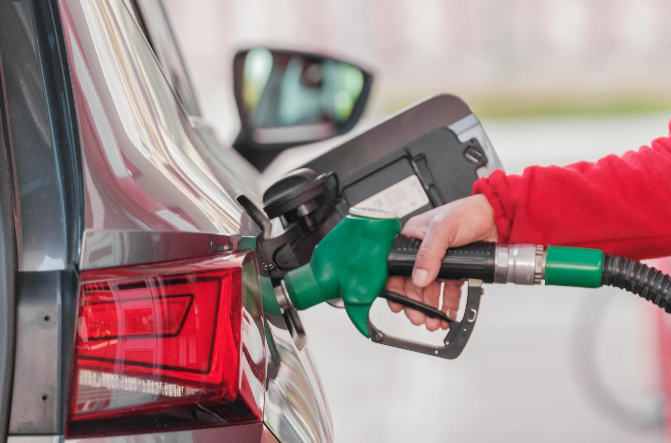 Taniej za benzynę i LPG dla dawców krwi. Jakie są zasady rabatu na stacjach? Ręka osoby ubrane w czerwoną bluzę trzyma zieloną rączkę dystrybutora paliwa i tankuje samochód osobowy, widoczny jest tylny reflektor oraz wlew paliwa.