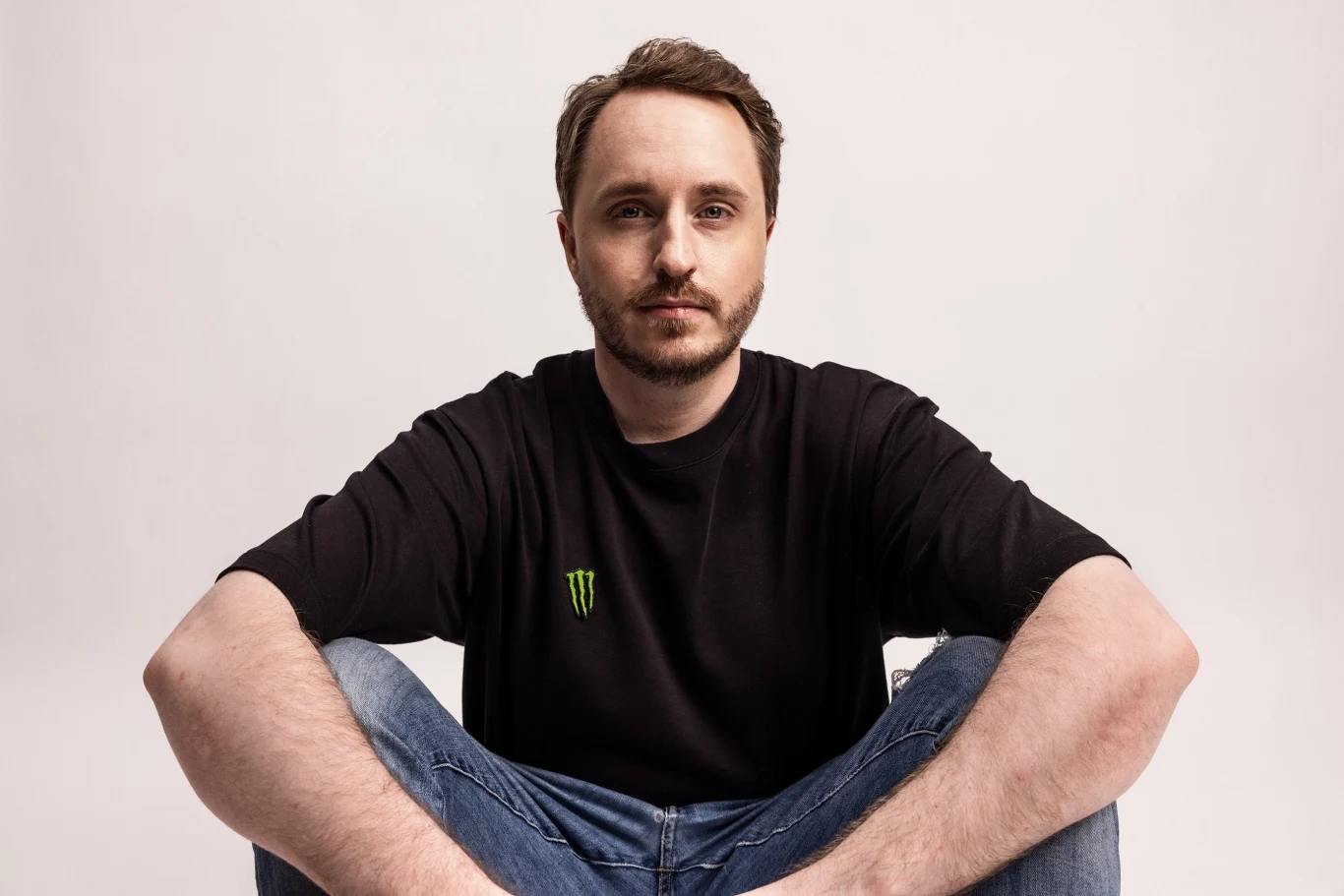GeT_RiGhT Mężczyzna z krótką brodą i jasnymi włosami siedzący ze skrzyżowanymi nogami, ubrany w czarną koszulkę z logo firmy Monster i niebieskie dżinsy, patrzy prosto w obiektyw na jasnym tle.