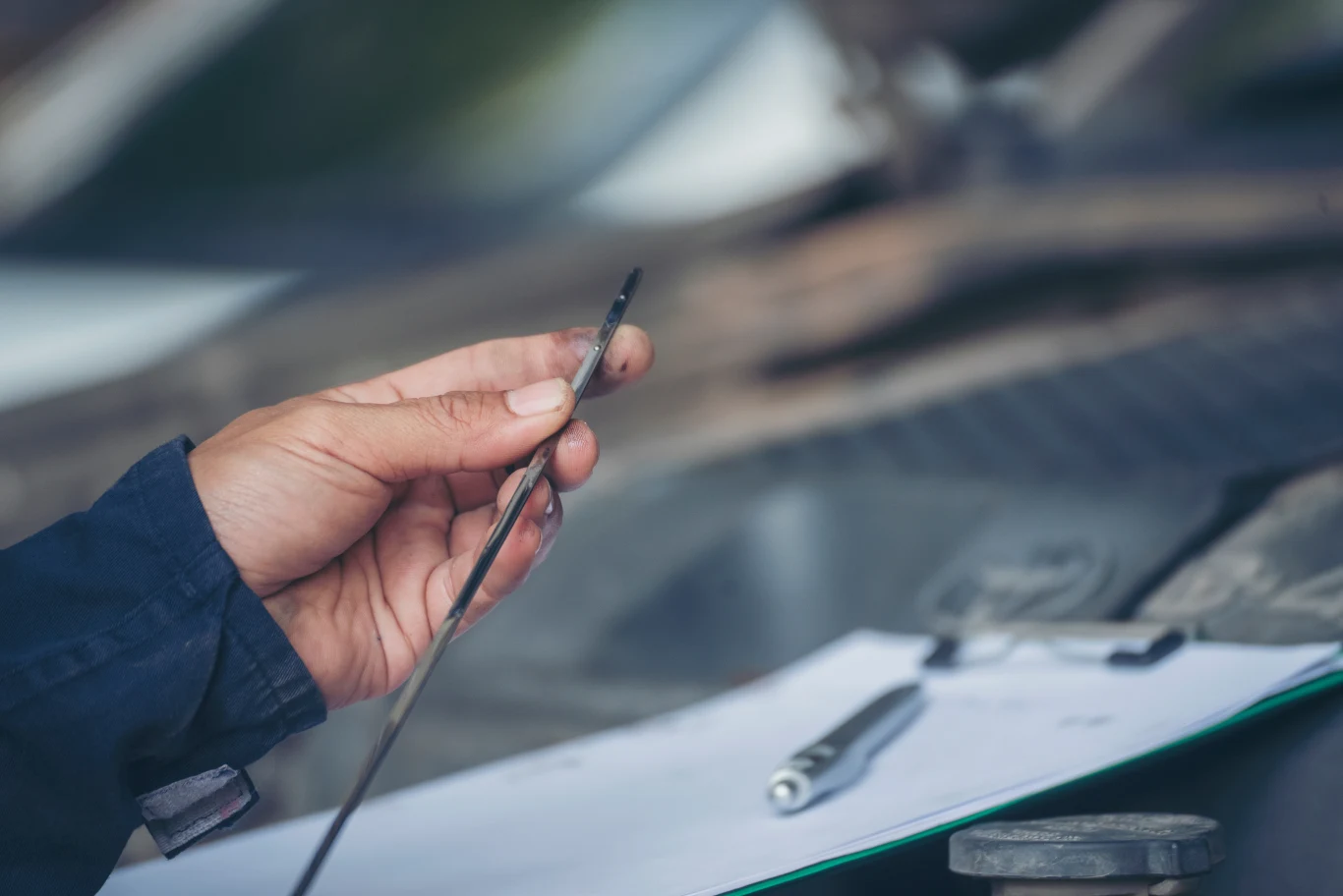 Każdy bagnet ma własny system oznakowania, ale podstawowa zasada pozostaje niezmieniona. Dłoń trzyma brudny bagnet do sprawdzania poziomu oleju silnikowego, w tle rozmazane narzędzia i bloczek z długopisem leżące na powierzchni przypominającej maskę samochodu.