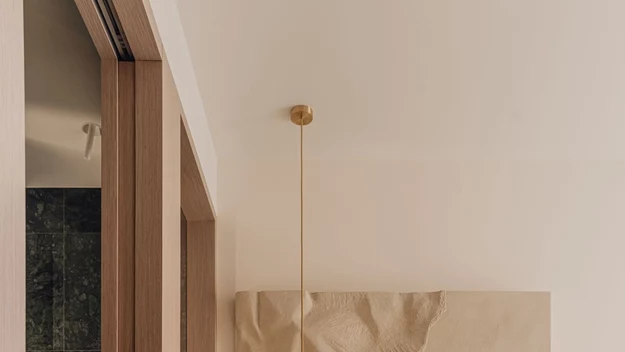 Minimalistyczna sypialnia z jasnymi ścianami, eleganckim łóżkiem z białą pościelą, nowoczesną lampą wiszącą oraz czarnym stolikiem nocnym, na którym stoi pudełko chusteczek; na ścianie nad łóżkiem dekoracyjny element artystyczny.