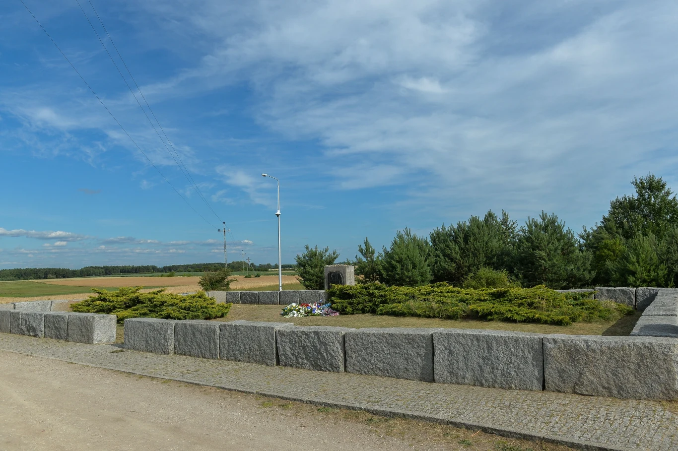 Pomnik w Jedwabnem Kamienny pomnik otoczony niskimi krzewami i wieńcami, w tle pola uprawne oraz niewielki las pod błękitnym niebem z lekkim zachmurzeniem.