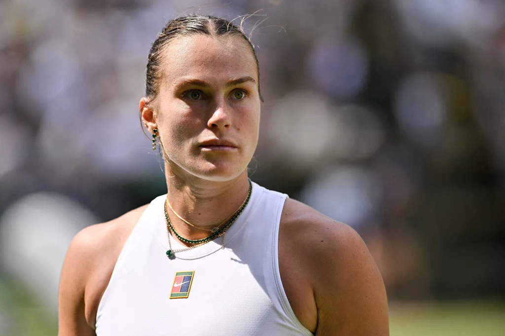 Aryna Sabalenka Kobieta w sportowym stroju stoi na tle rozmytego stadionu, z wyrazem skupienia na twarzy, nosi kilka naszyjników i sportową koszulkę bez rękawów.
