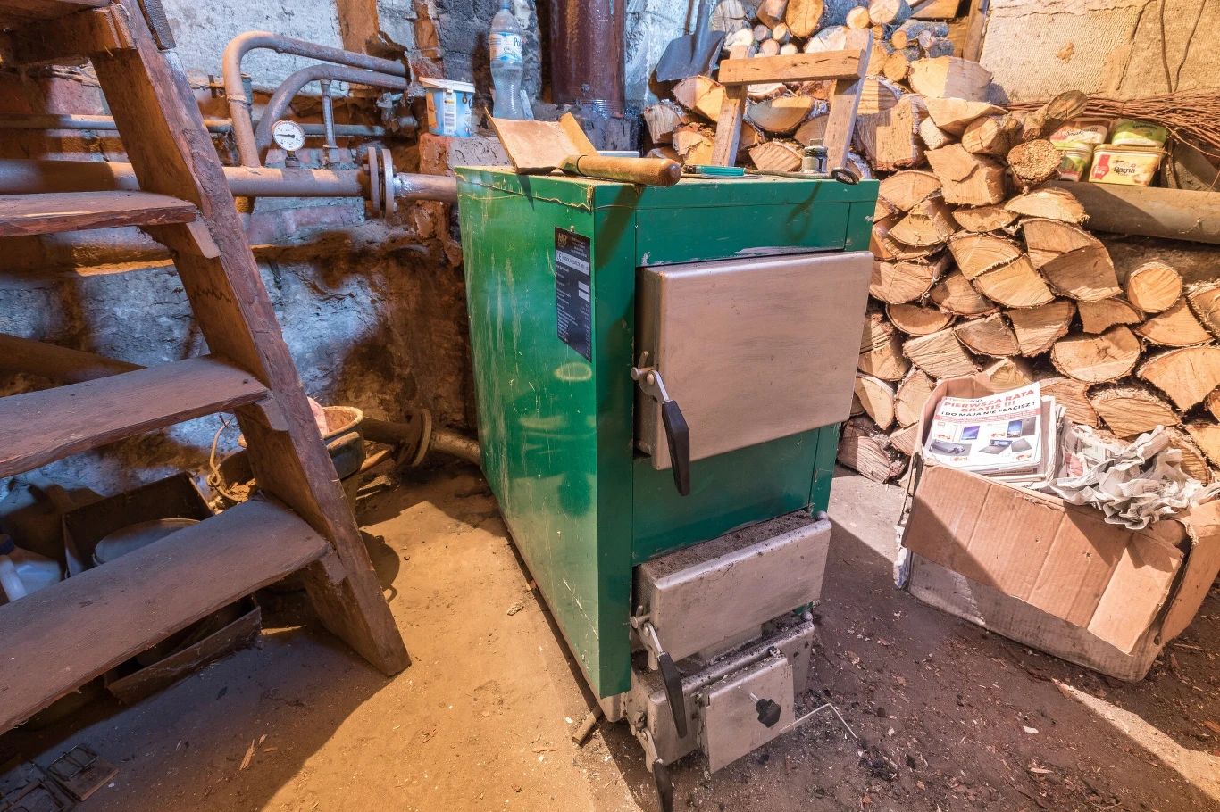 Niektórzy beneficjenci programu "Czyste Powietrze" po kontrolach muszą płacić zaliczki, mimo że nie wymieniono im pieców Piec centralnego ogrzewania w zielonej obudowie ustawiony w piwnicy obok stosu drewna opałowego oraz kartonu z gazetami, po lewej stronie drewniane schody prowadzące na wyższy poziom.