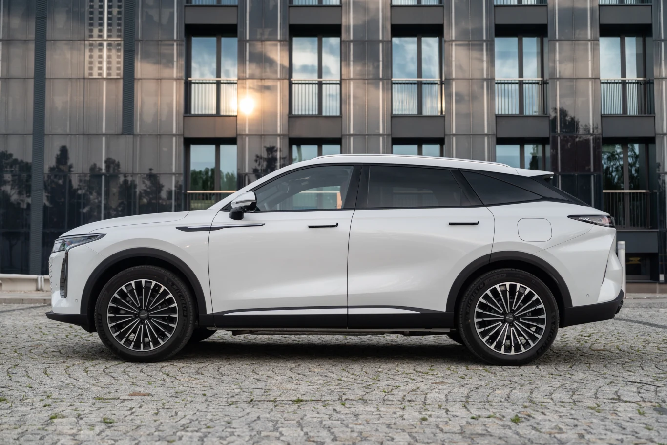 Biały nowoczesny SUV zaparkowany na brukowanym placu na tle przeszklonego budynku o regularnych oknach, wyraźne linie nadwozia i duże felgi podkreślają dynamiczny charakter samochodu.