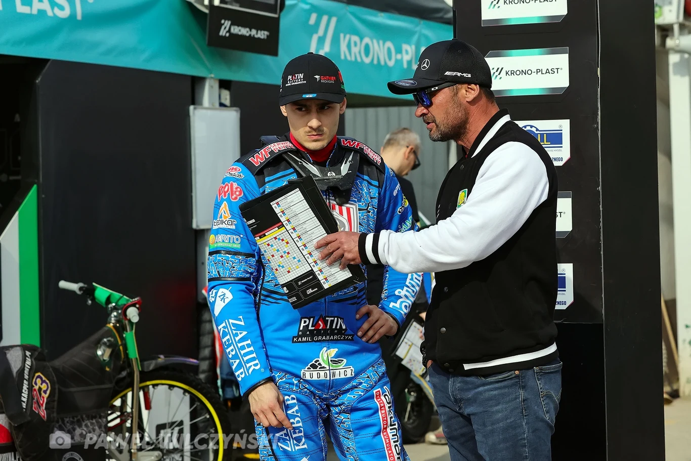 Dwóch mężczyzn w sportowej strefie serwisowej, jeden ubrany w strój żużlowca z licznymi logotypami sponsorów, drugi w czapce i okularach przeciwsłonecznych omawiający strategię z pomocą planszy z rozkładem biegów; w tle motocykl żużlowy.