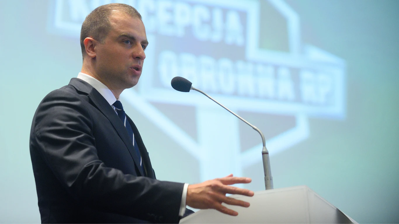 Tomasz Szatkowski Mężczyzna w garniturze przemawiający przy mównicy, w tle rozmyślony ekran z napisem i symbolem graficznym, prawdopodobnie w trakcie oficjalnej prezentacji lub konferencji.