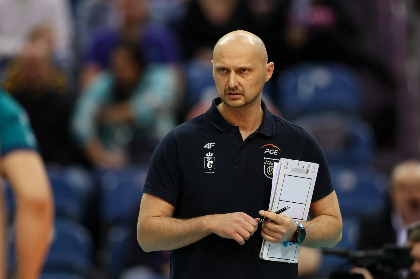 Mężczyzna w sportowej koszulce z emblematami klubu siatkarskiego trzymający w ręku tabliczkę z kartkami, w tle rozmyci kibice na trybunach.