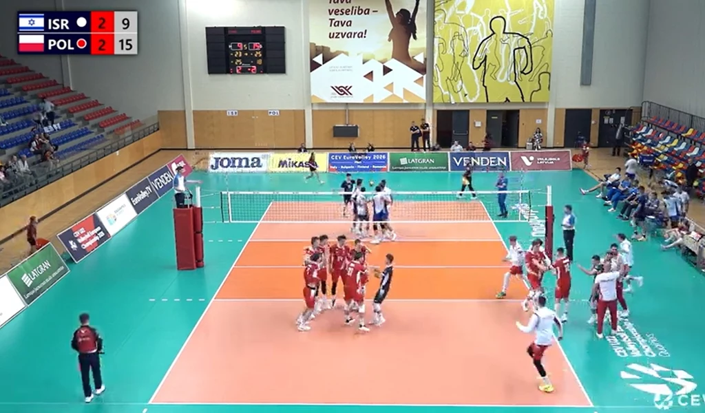 Radość Polaków po ostatniej piłce w meczu z Izraelem Mecz siatkówki halowej z udziałem dwóch drużyn narodowych w sali sportowej, na tablicy wyników przewaga Polski nad Izraelem, po środku kortu skupieni zawodnicy, wokół trybuny z niewielką liczbą widzów i ławki rezerwowych.