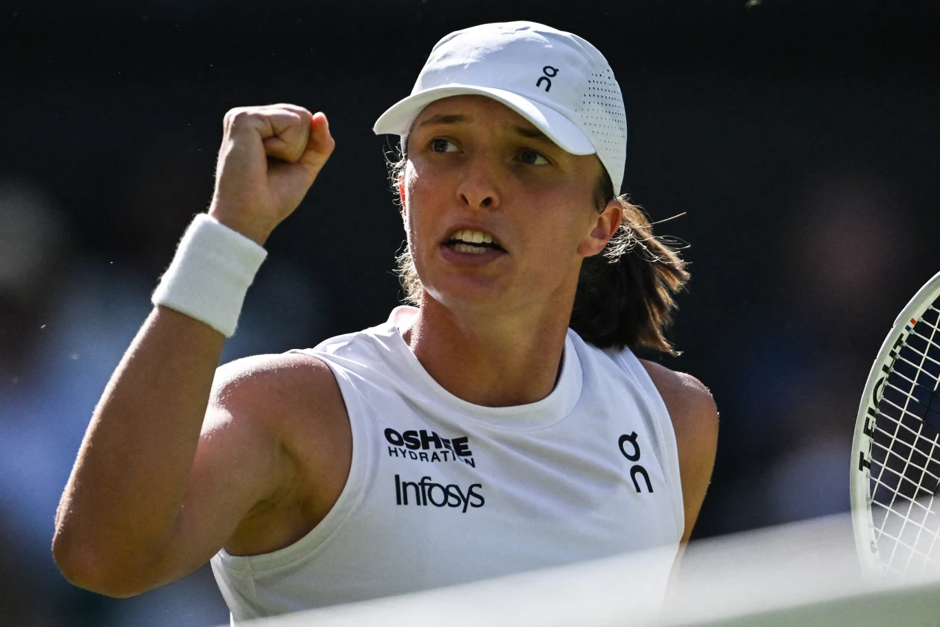 Iga Świątek rywalizowała z Belindą Bencić o awans do finału Wimbledonu Tenisistka w białym stroju i czapce z logo, trzymająca rakietę i zaciśniętą pięścią, wyrażająca emocje na korcie podczas meczu tenisowego.