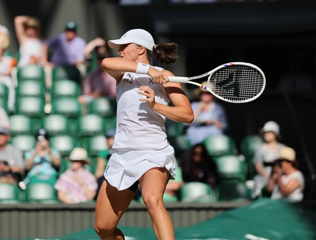 Iga Świątek, Wimbledon 2025 Tenisistka w białym stroju wykonująca zamach rakietą na korcie, w tle widać trybuny z siedzącymi widzami.