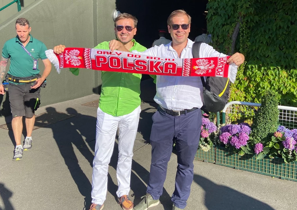 Dwóch mężczyzn stojących obok siebie trzyma rozwinięty szalik kibica z napisem 'Polska' oraz orłami, uśmiechają się i pozują do zdjęcia na tle zieleni i kwiatów, w tle spacerująca osoba.