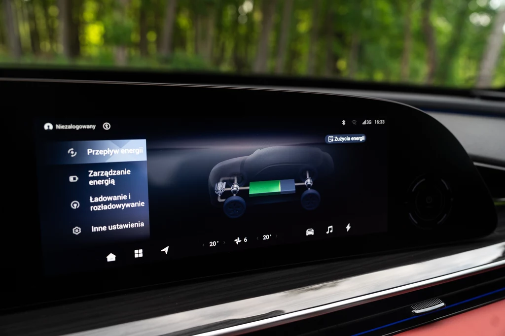 Omoda 9 pozwala na przejechanie naprawdę dużego dystansu na prądzie - w teście było to 168 km Wnętrze samochodu elektrycznego z wyświetlaczem prezentującym menu zarządzania energią oraz schemat przepływu energii między baterią a kołami pojazdu, na tle rozmytego lasu widocznego przez szybę.