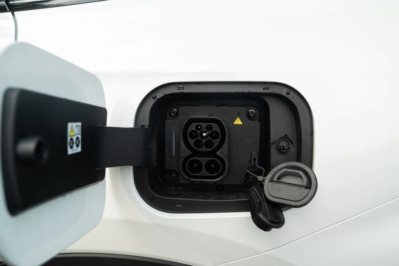 Gniazdo ładowania samochodu elektrycznego z widocznym kablem oraz klapką ochronną na białej karoserii pojazdu elektrycznego.