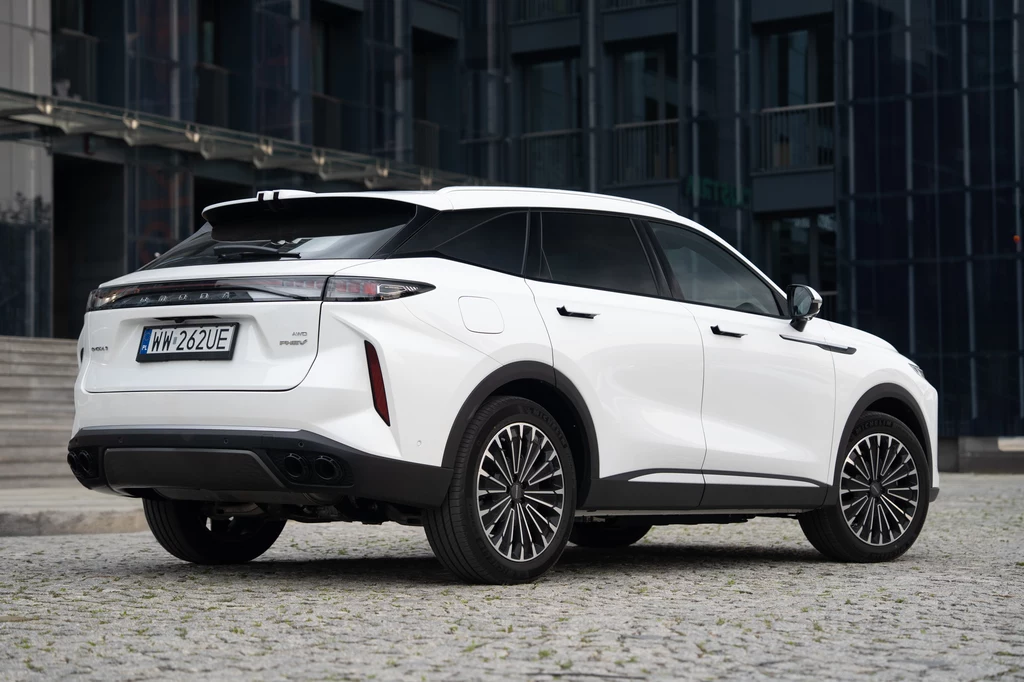 Stylistyka Omody 9 jest wyraźnie azjatycka, ale bez wątpienia skrojona pod europejskie gusta Biały samochód osobowy typu SUV z przyciemnianymi szybami stoi na brukowanym placu przed nowoczesnym, szklanym budynkiem, widoczne są detale tylnej części pojazdu oraz jego tablica rejestracyjna.