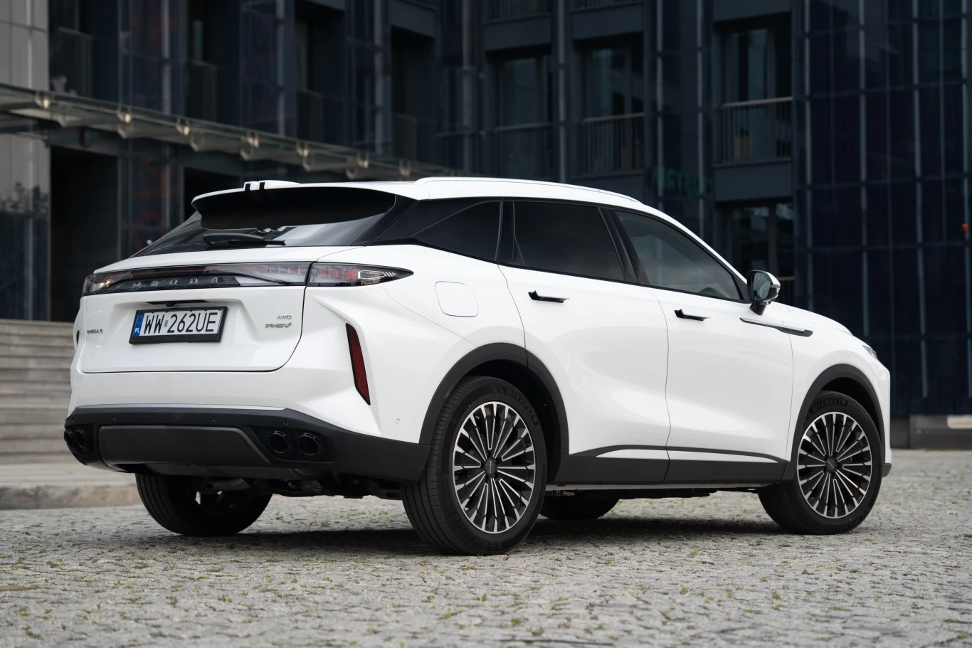 Biały samochód osobowy typu SUV z przyciemnianymi szybami stoi na brukowanym placu przed nowoczesnym, szklanym budynkiem, widoczne są detale tylnej części pojazdu oraz jego tablica rejestracyjna.