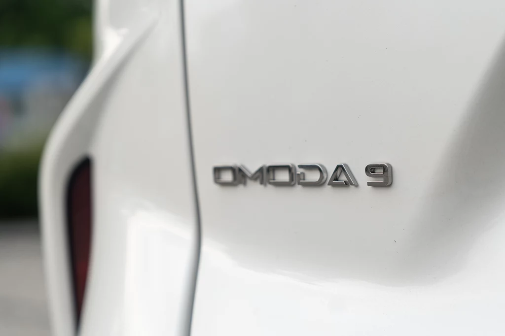 Omoda 9 Białe nadwozie samochodu z wyraźnie widocznym emblematem Omoda 9 po prawej stronie kadru.