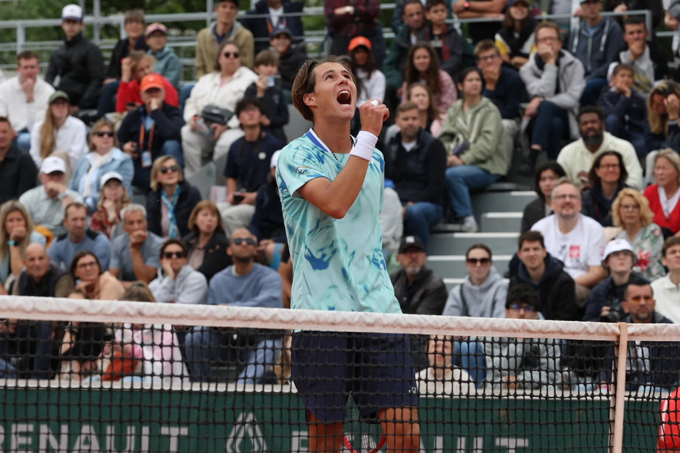 Alan Ważny odpadł z singla, ale wciąż pozostaje w grze o tytuł w deblu na juniorskim Wimbledonie Tenisista w błękitnej koszulce wyraża emocje po zdobyciu punktu, stoi przy siatce na korcie, w tle liczna publiczność obserwująca mecz.