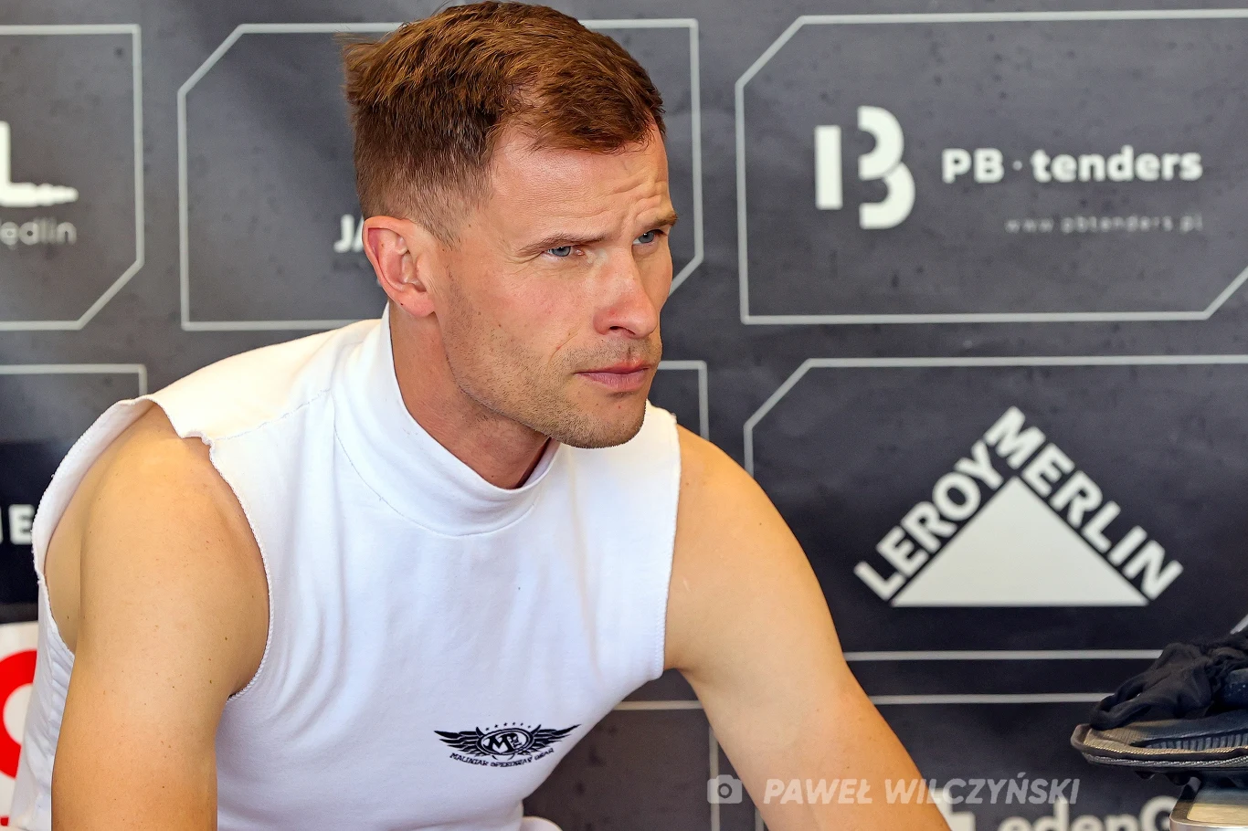 Matej Zagar Mężczyzna w sportowej koszulce bez rękawów siedzi na tle czarnej ściany z logo różnych marek i sponsorów, skupiony wzrok skierowany lekko w bok.