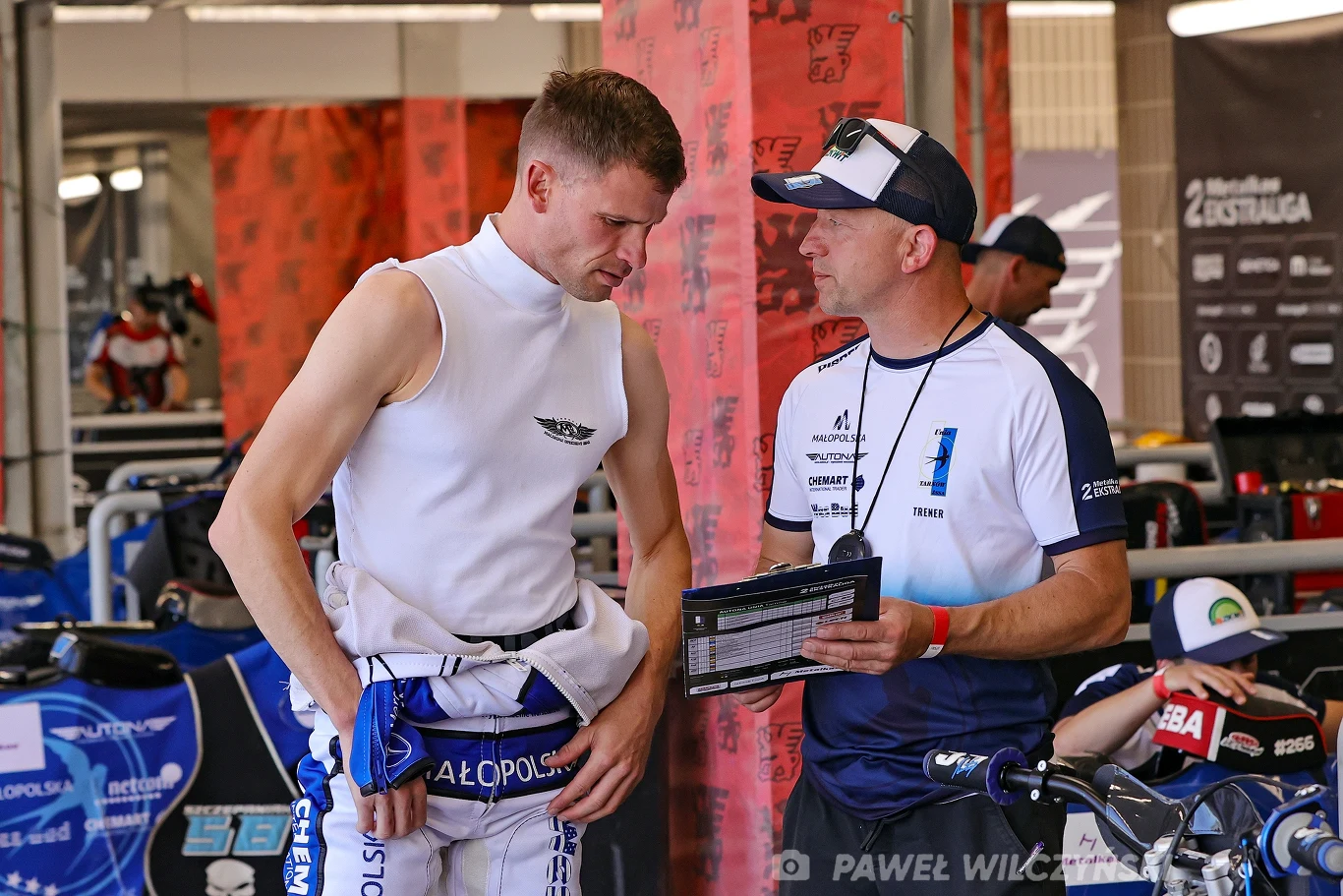 Matej Zagar i Stanisław Burza Dwóch mężczyzn rozmawia w garażu sportowym, jeden z nich ubrany w strój żużlowy, prawdopodobnie zawodnik, drugi trzyma notatki lub tablet, obaj skoncentrowani na omawianiu strategii lub ustawień motocykla. W tle widać plandeki i elementy wyposażenia ze...