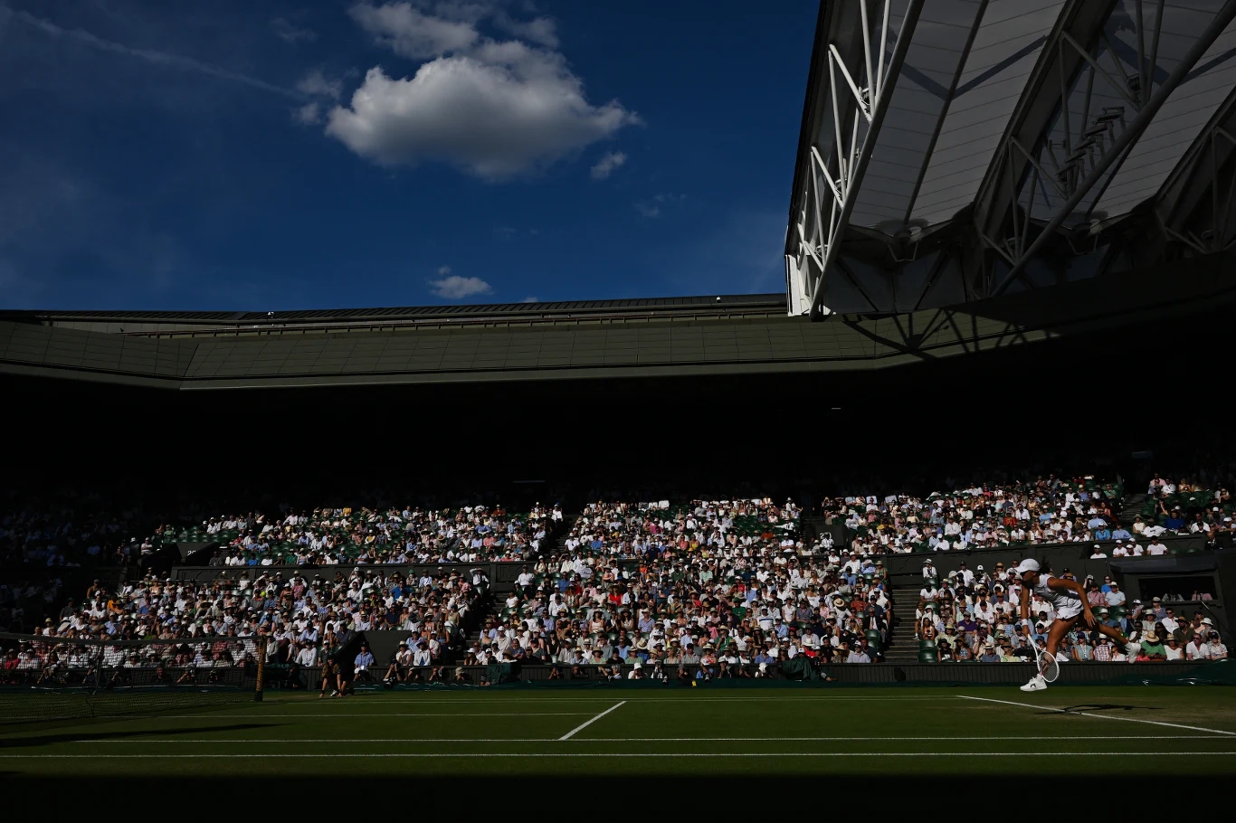 Kort centralny Wimbledonu Kort tenisowy podczas meczu, zawodnik serwuje piłkę, liczna publiczność obserwuje wydarzenie z trybun a jasne światło dzienne pada na fragment boiska, reszta otoczenia pozostaje w cieniu.