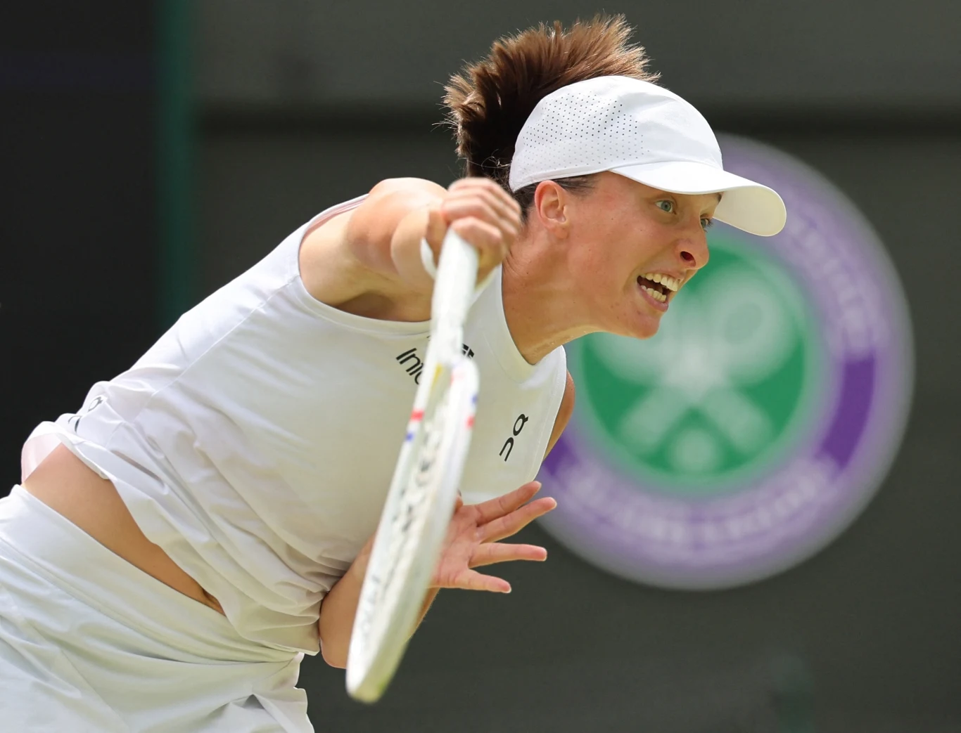Zawodniczka w białym stroju sportowym wykonuje dynamiczny serwis podczas meczu tenisowego na trawiastym korcie, w tle widać rozmazany symbol turnieju tenisowego Wimbledon.