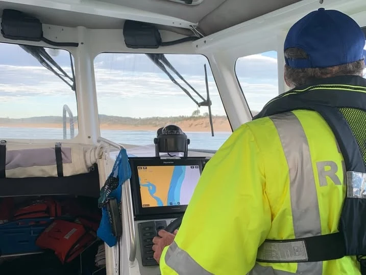 Nastoletni surfer odnalazł się na bezludnej wyspie Operator łodzi motorowej w jaskrawej, odblaskowej kurtce i czapce steruje pojazdem na wodzie, korzystając z nowoczesnej nawigacji elektronicznej; przez okna widoczny fragment lądu oraz spokojna tafla wody.