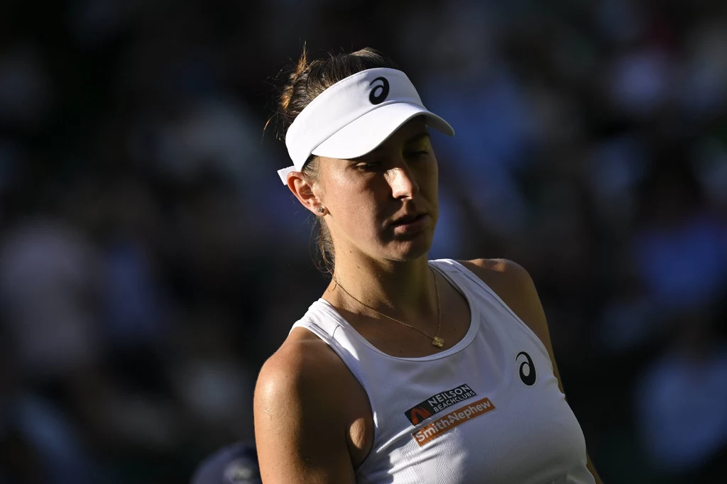 Belinda Bencic Kobieta w białym stroju sportowym i białej czapce z daszkiem stoi na tle rozmytego tła, na jej koszulce widoczny jest logotyp sponsora oraz symbol producenta odzieży sportowej, twarz przysłonięta częściowo cieniem.