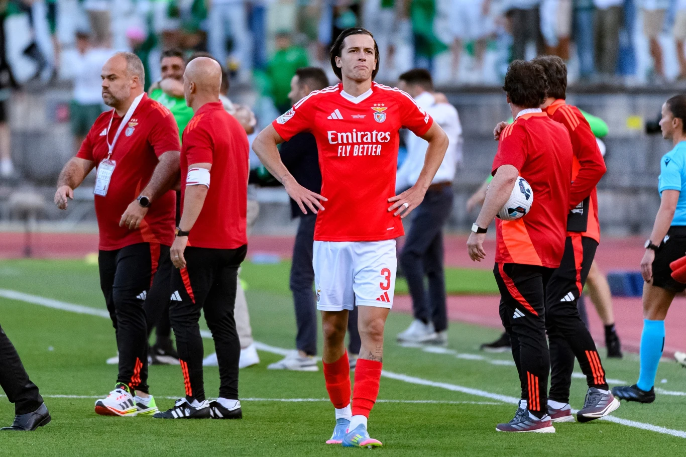 Alvaro Carreras Piłkarz drużyny Benfica w czerwonym stroju stoi na murawie z rękami na biodrach, wokół znajdują się członkowie sztabu trenerskiego i inni uczestnicy wydarzenia, w tle publiczność na trybunach.