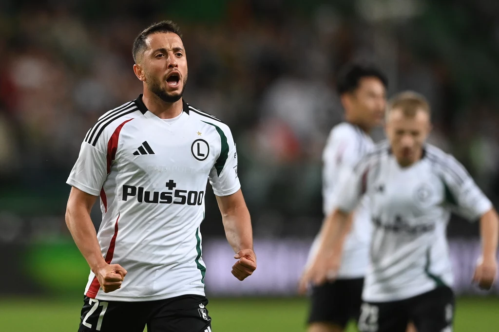 Wahan Biczachczjan tuż po zdobyciu bramki na 1:0 Piłkarz drużyny Legia Warszawa w białym stroju z logo sponsora okazuje wybuch emocji, zaciskając pięści i krzycząc po zdobyciu gola na tle rozmytych sylwetek innych piłkarzy.