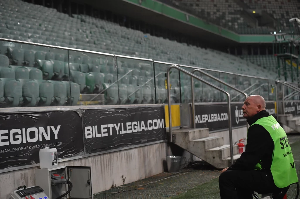 "Żyleta"... cicha jak nigdy Ochroniarz w zielonej kamizelce siedzi przy pustych trybunach stadionu, na barierkach widoczne są banery reklamowe z napisem bilety.legia.com.