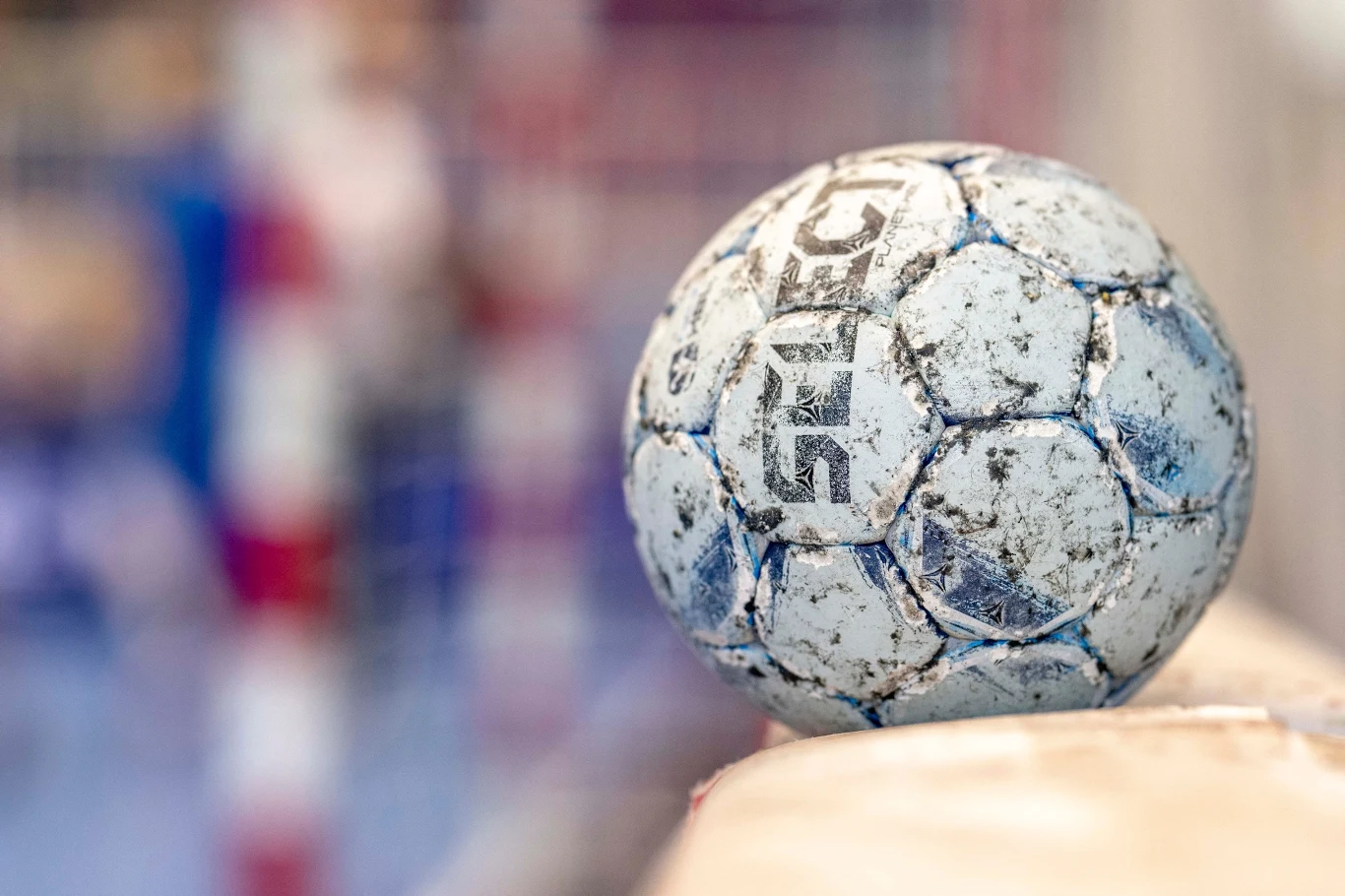 Zużyta piłka ręczna oparta na krawędzi ławki lub bandy, w tle rozmyta bramka ozdobiona czerwono-białymi słupkami.