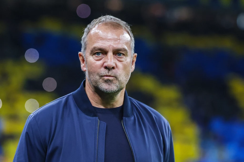 Hansi Flick Mężczyzna w średnim wieku z krótkimi, siwiejącymi włosami ubrany w granatową kurtkę na tle rozmytego stadionu z żółto-niebieskimi krzesełkami.