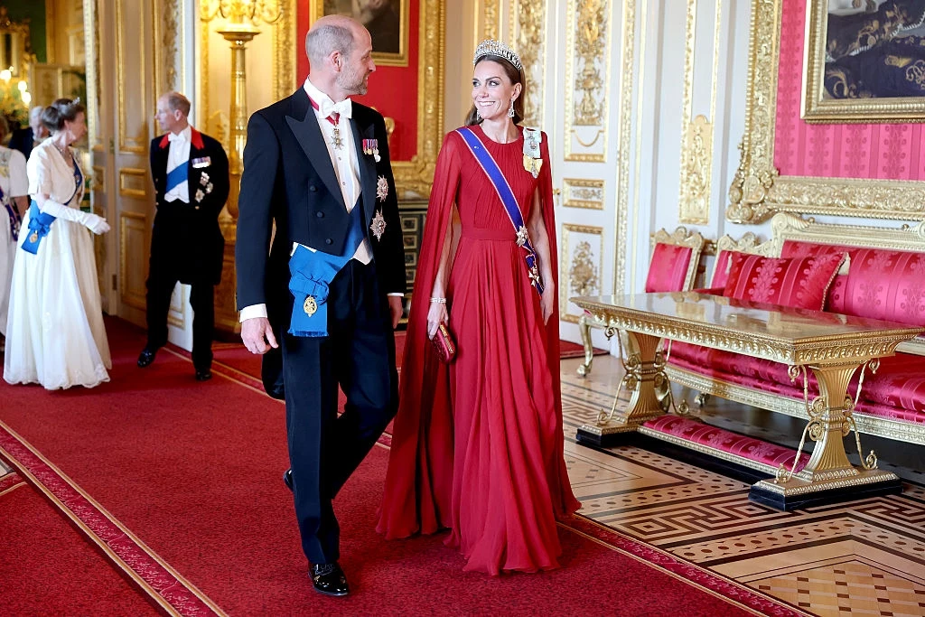 Kreacja księżnej Kate podczas bankietu Para ubrana w eleganckie stroje wieczorowe idzie po czerwonym dywanie w bogato zdobionym pałacowym wnętrzu z pozłacanymi meblami i czerwonymi fotelami, kobieta ma czerwoną suknię i tiarę.