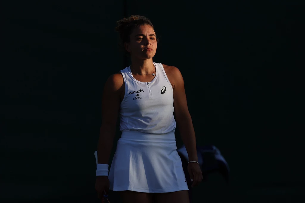 Jasmine Paolini Tenisistka ubrana w biały sportowy strój stoi na korcie, silnie oświetlona przez słońce na ciemnym tle, z wyrazem skupienia i determinacji na twarzy.