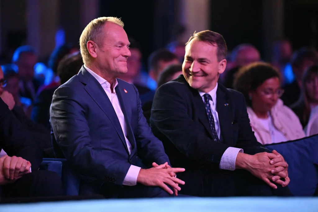 Dwóch mężczyzn w garniturach siedzi w pierwszym rzędzie podczas wydarzenia, uśmiechają się i rozmawiają, w tle widać innych uczestników spotkania.