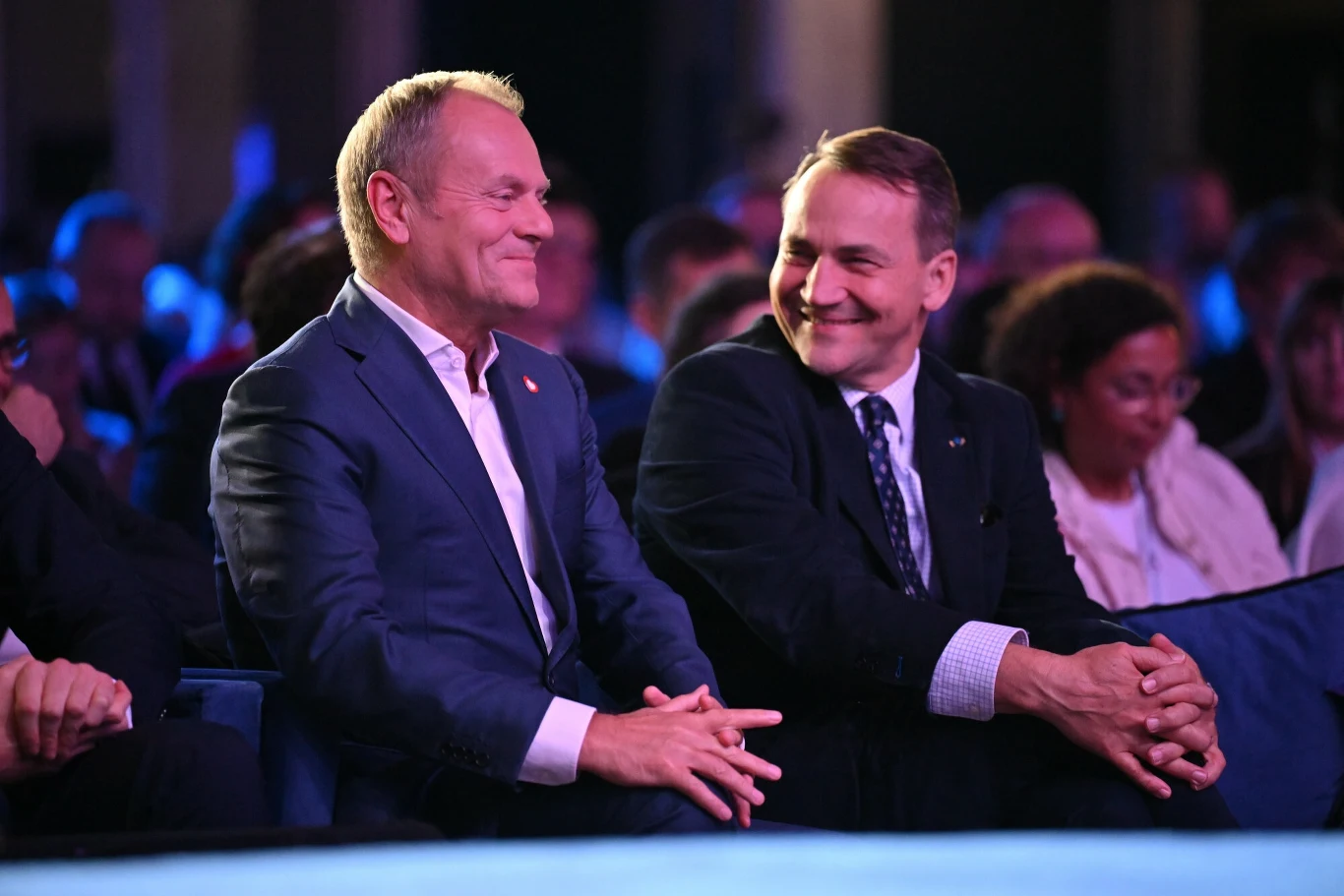 Dwóch mężczyzn w garniturach siedzi w pierwszym rzędzie podczas wydarzenia, uśmiechają się i rozmawiają, w tle widać innych uczestników spotkania.